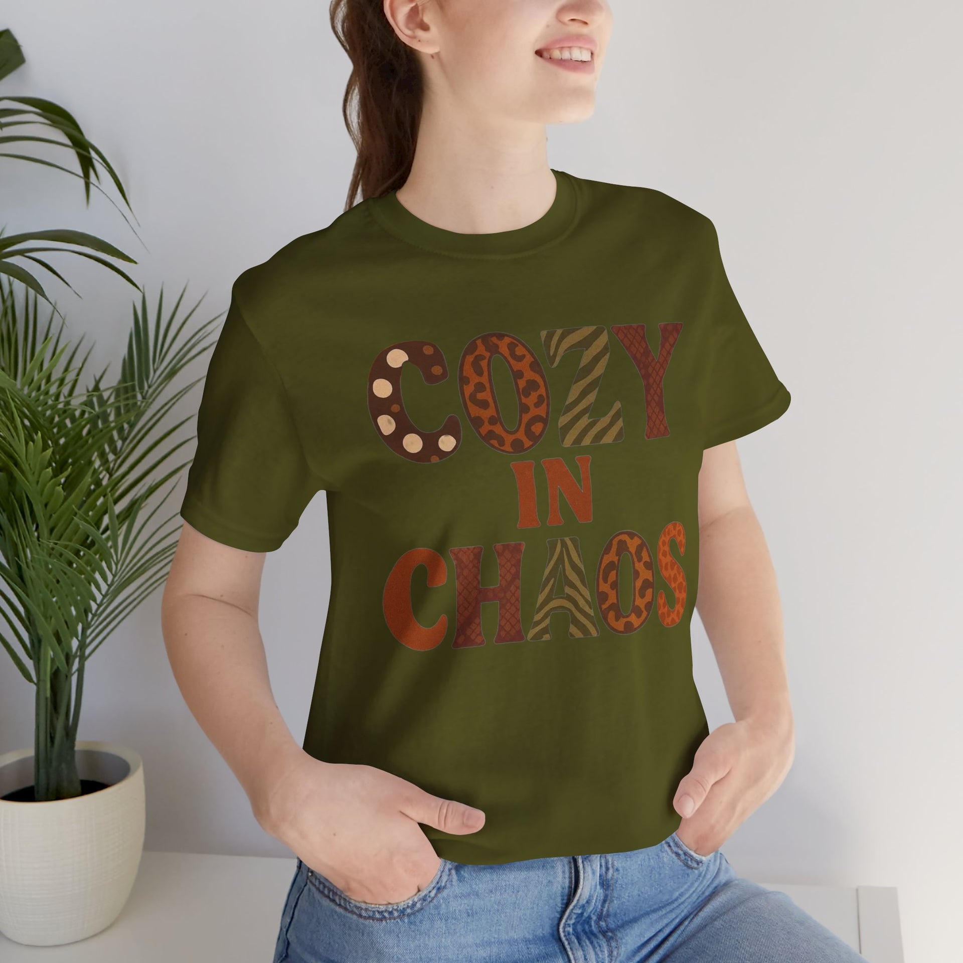 Person wearing a green t-shirt with 'COZY IN CHAOS' text, standing indoors next to a plant.