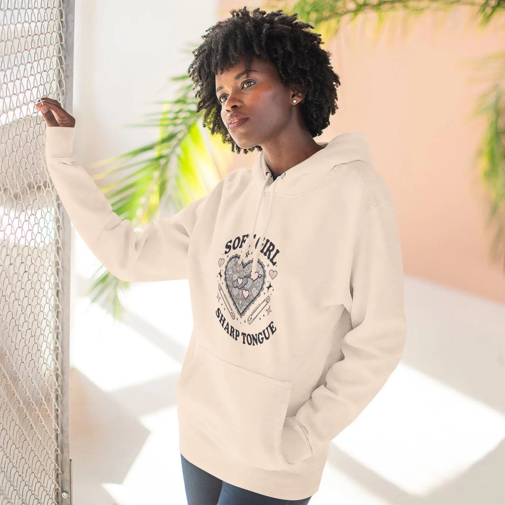 Woman wearing a beige hoodie with text and graphics, standing against a light background with plants.