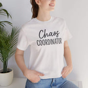 Person wearing a white t-shirt with 'Chaos Coordinator' text, standing indoors next to a plant.