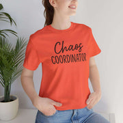 Person wearing an orange t-shirt with 'Chaos Coordinator' text, standing indoors next to a plant.