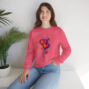 Person wearing a pink sweatshirt with a colorful heart design, sitting against a white wall with a plant to the left.
