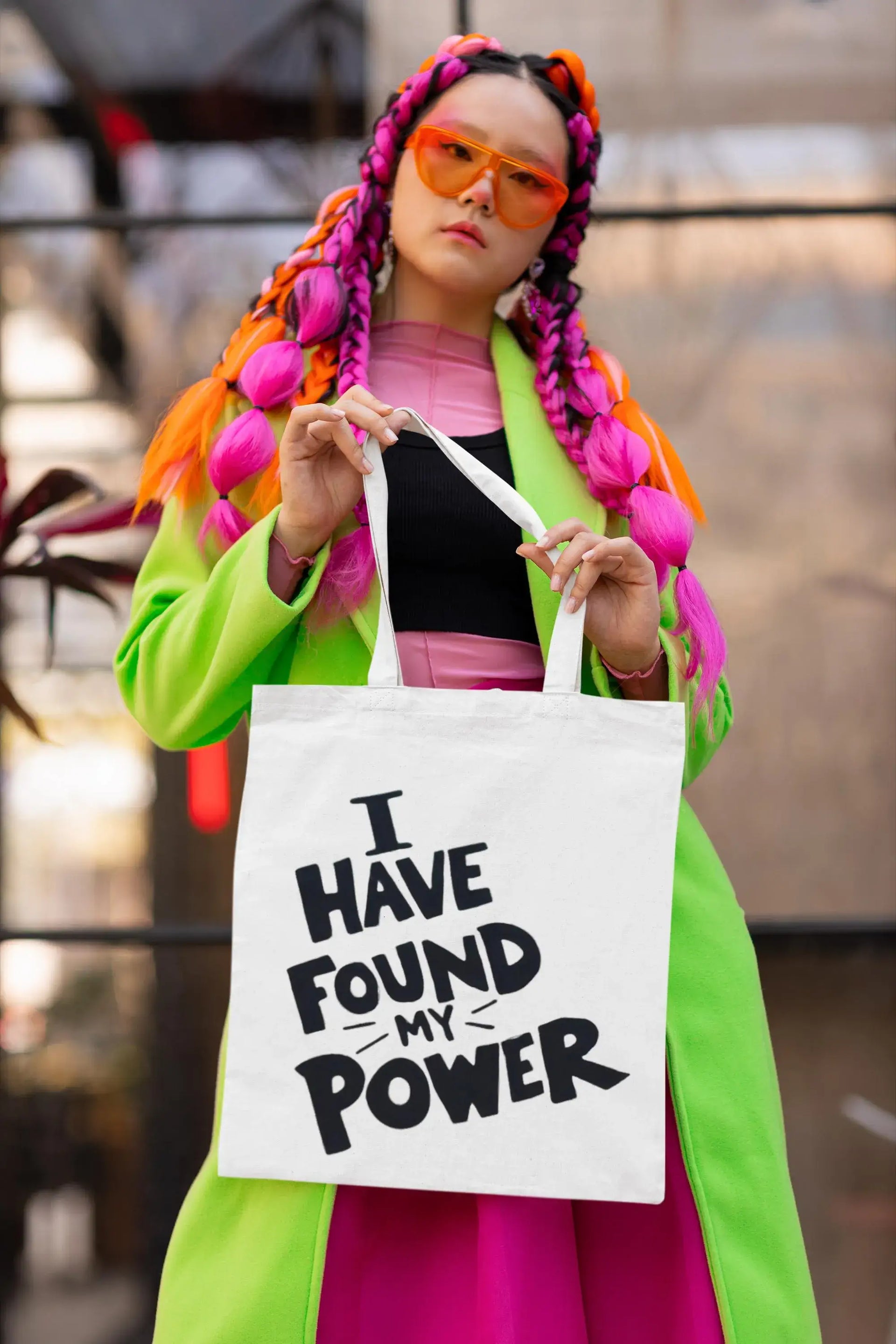 Person with colorful hair and sunglasses holding a tote bag with text, blurred background