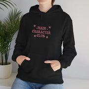 Person wearing a black hoodie with 'Main Character Club' text and stars, standing indoors with a plant in the background.