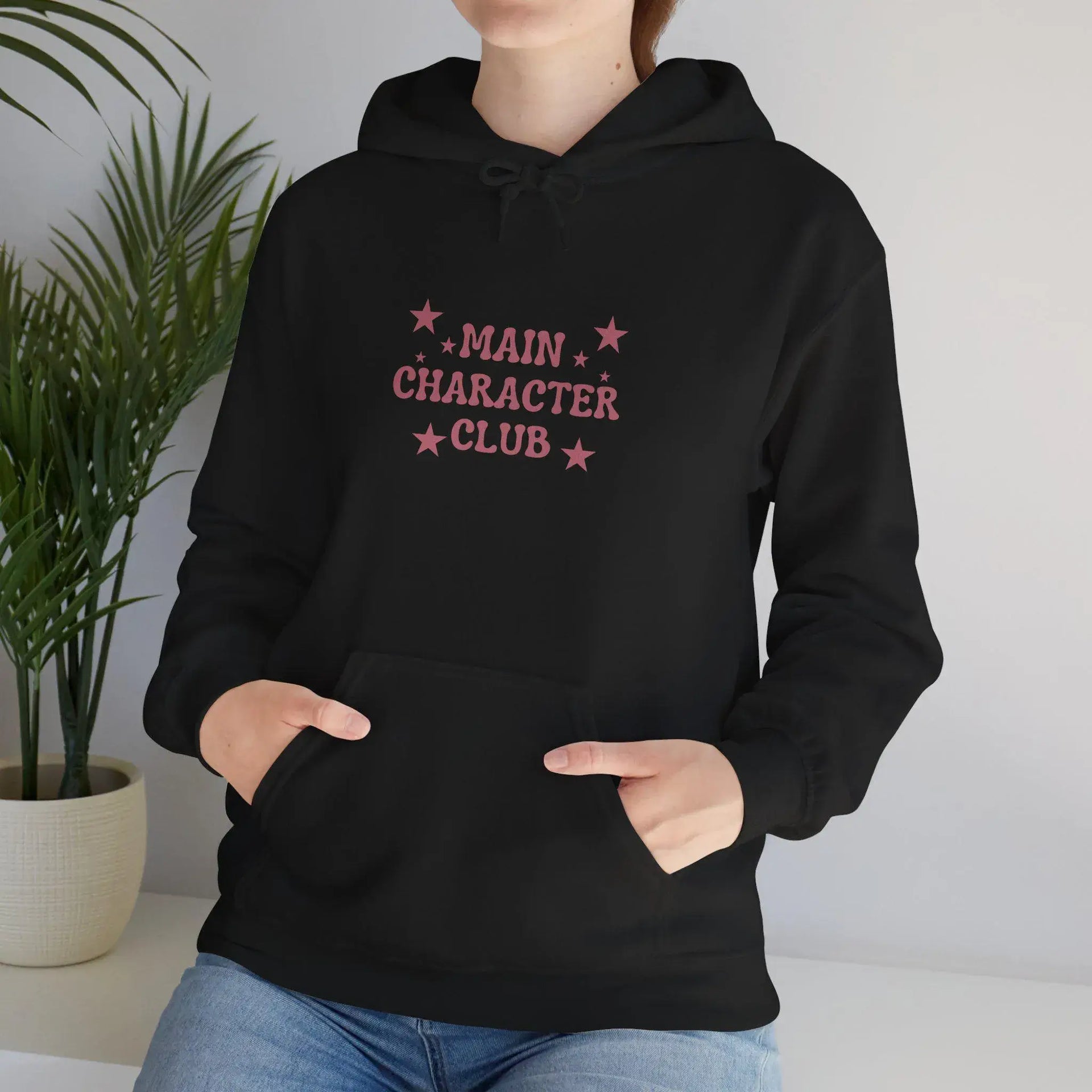 Person wearing a black hoodie with 'Main Character Club' text and stars, standing indoors with a plant in the background.