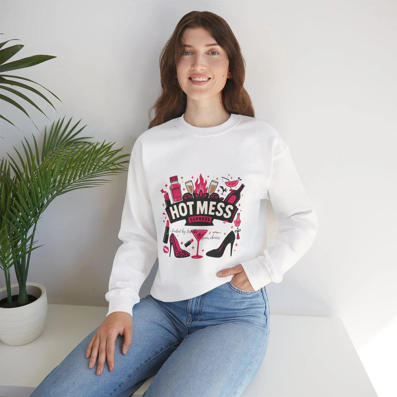 Woman wearing a white sweatshirt with a graphic design sitting on a white surface with a plant in the background.